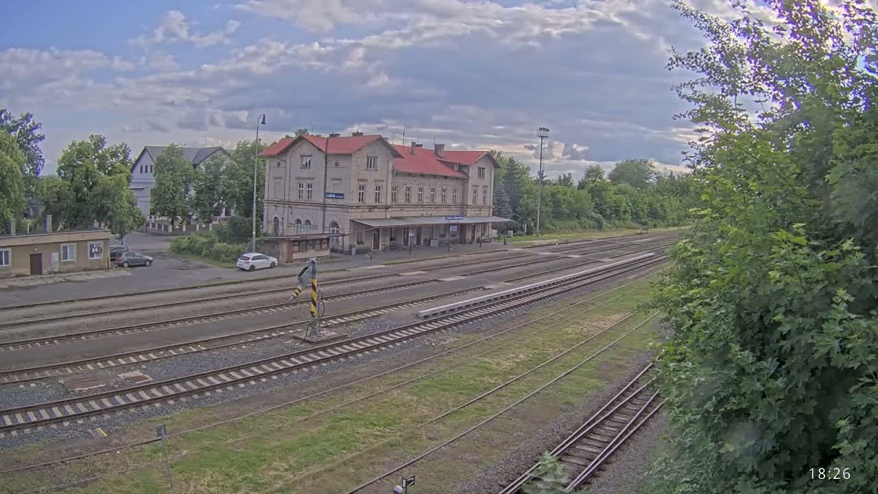 A partly cloudy day shows a train station with multiple tracks and a large building, surrounded by greenery.