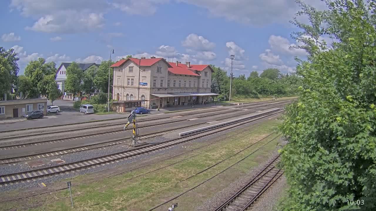 A train station with a red-roofed building sits beside multiple train tracks on a partly cloudy day.