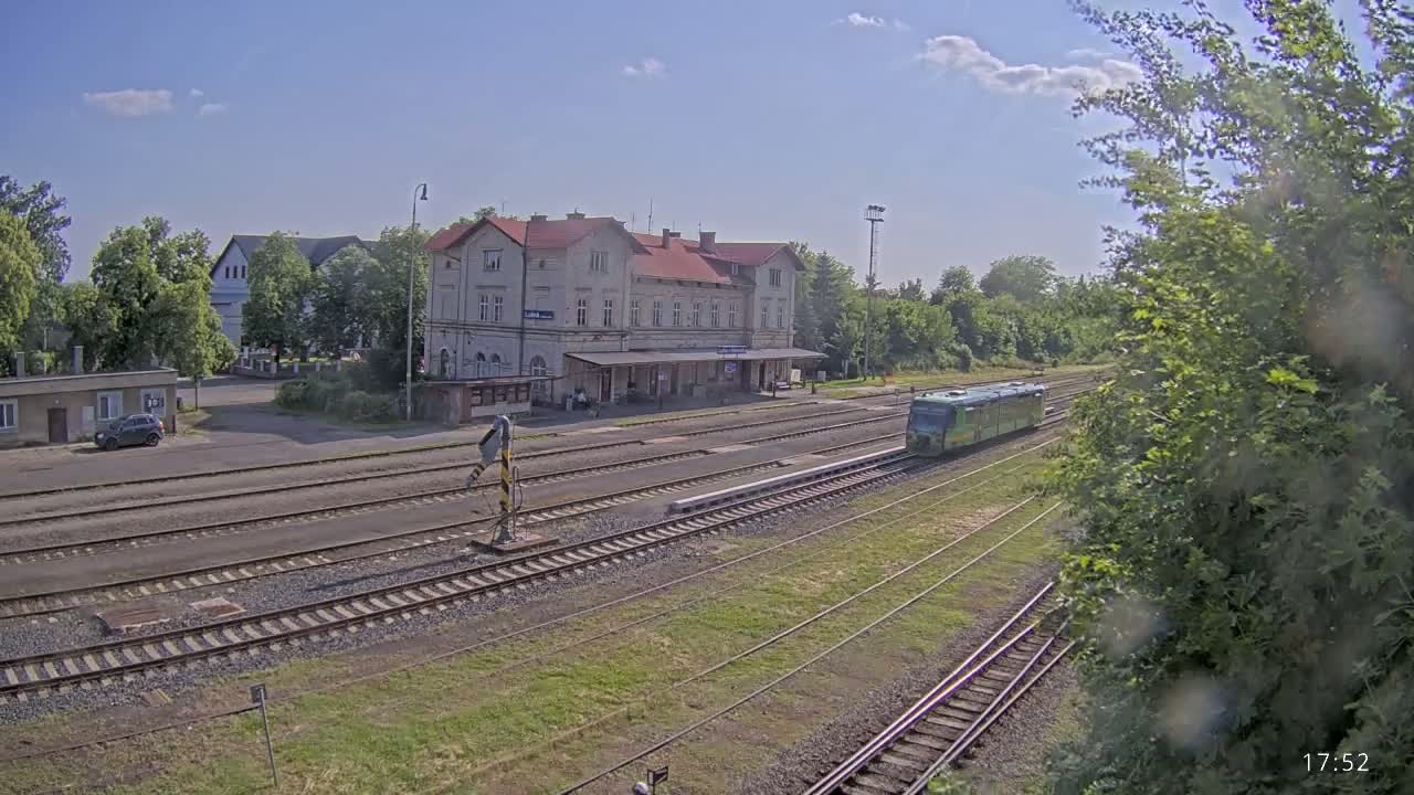 A green train is departing from a train station on a sunny day, with several tracks leading away from a long, light-colored building.