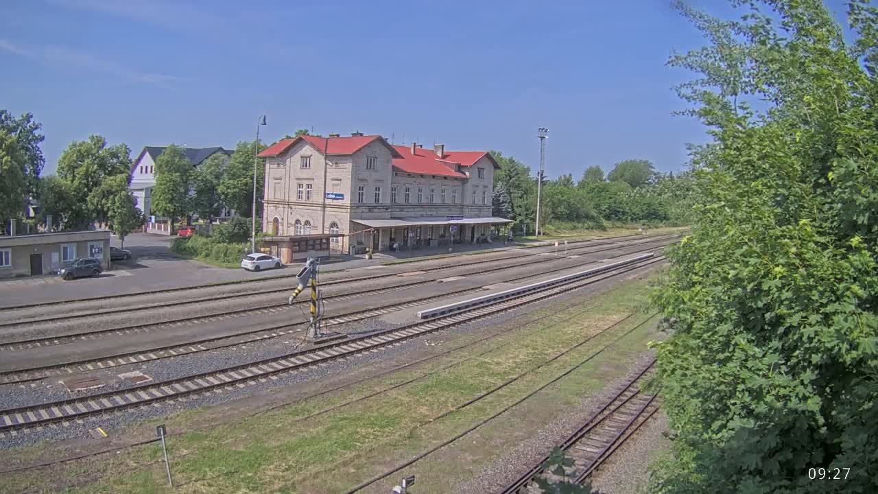 A train station building with a red roof sits beside several train tracks on a sunny day.
