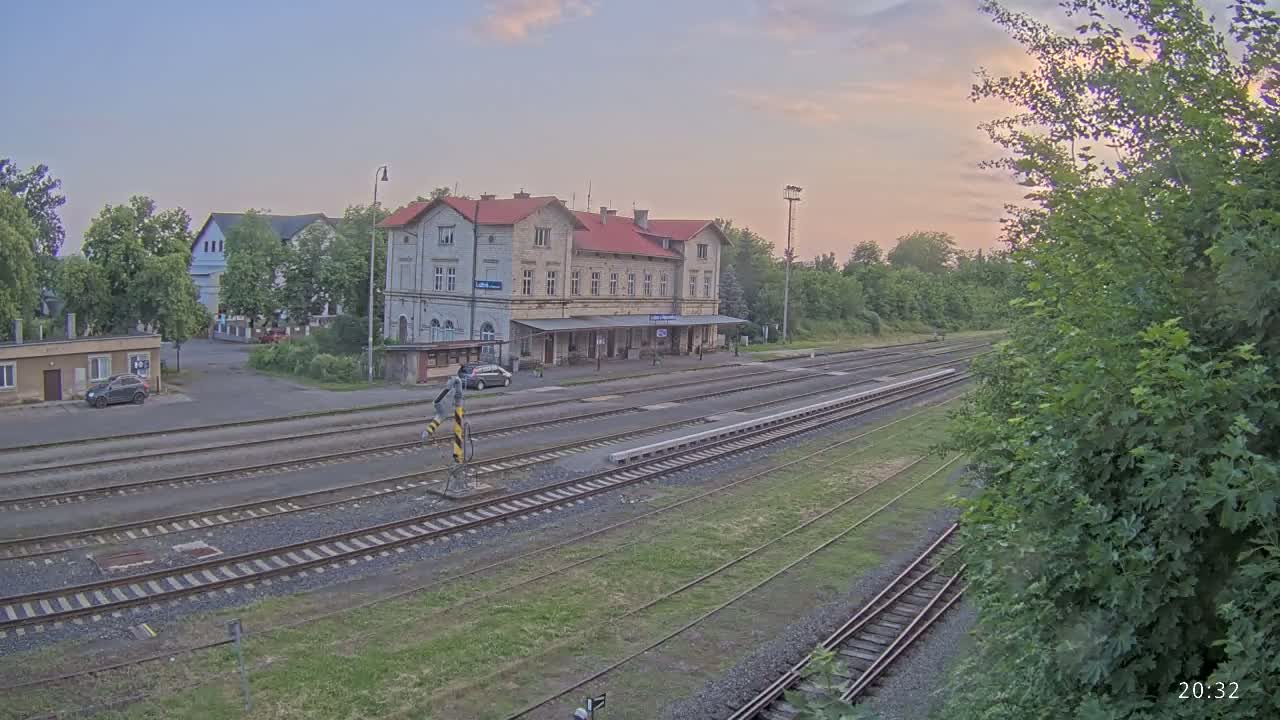 A train station with a long platform and several train tracks is visible under a partly cloudy, dusky sky.