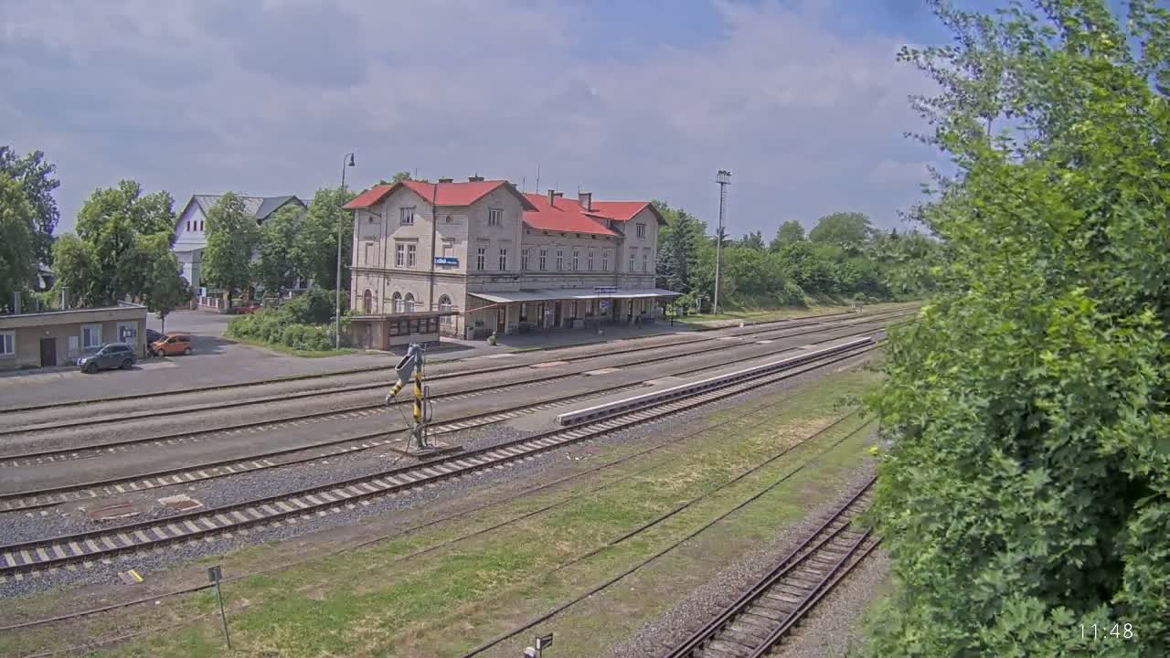 A long-shot view of a train station building with a red roof and several railway tracks extending into the distance on a partly cloudy day.