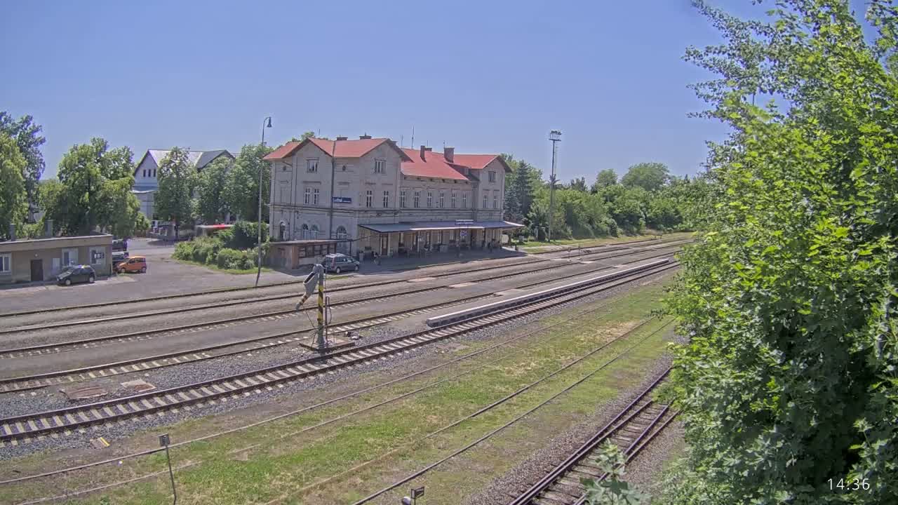 A train station with a long platform and multiple train tracks is seen on a sunny day, surrounded by green trees and other buildings.