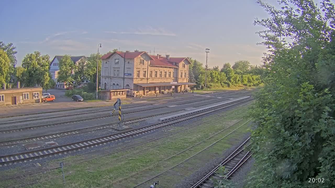A train station building with a red roof sits beside several railway tracks on a sunny day.