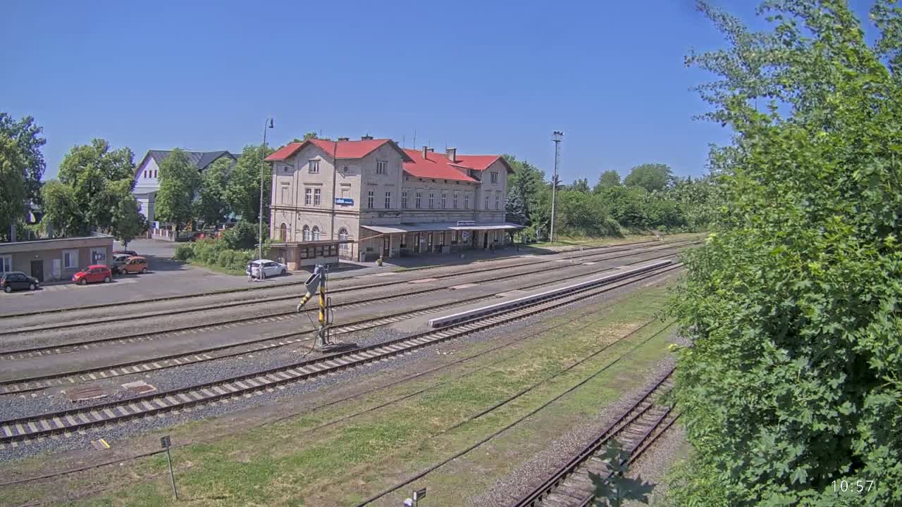A long train station building with a red roof sits beside several train tracks on a sunny day.