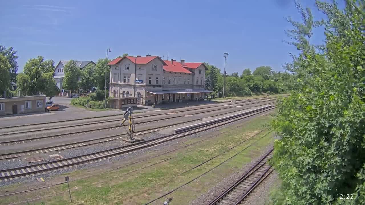 A train station with a long platform and several train tracks is situated in a sunny area surrounded by trees and other buildings.