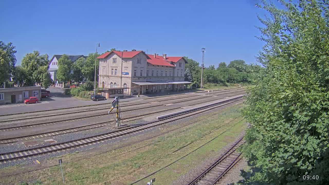 A train station with a long platform and several tracks is visible on a sunny day.