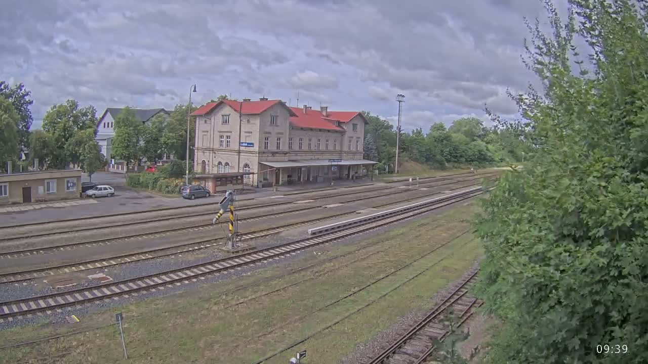 A train station with a long platform and several tracks is visible under a cloudy sky, with a few cars parked nearby and lush green vegetation surrounding the area.