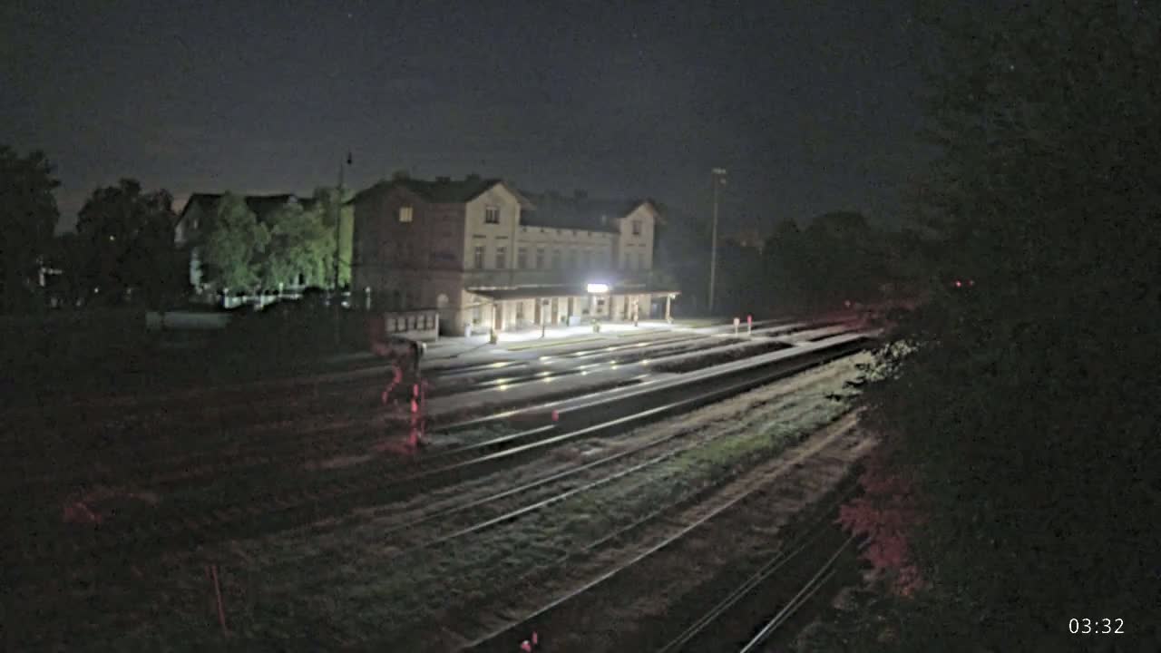 A train station with a long platform and several tracks is visible under a cloudy sky, with a few cars parked nearby and lush green vegetation surrounding the area.
