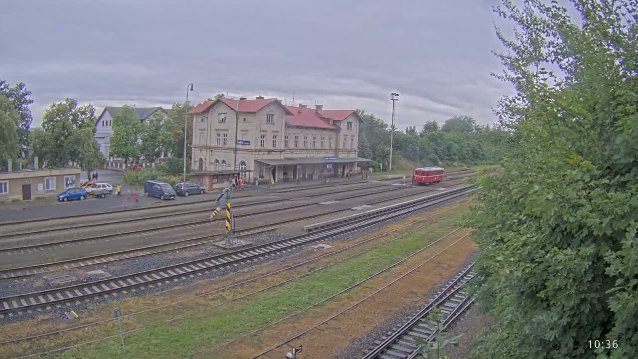 A train station with a red railcar and several cars parked nearby under an overcast sky.