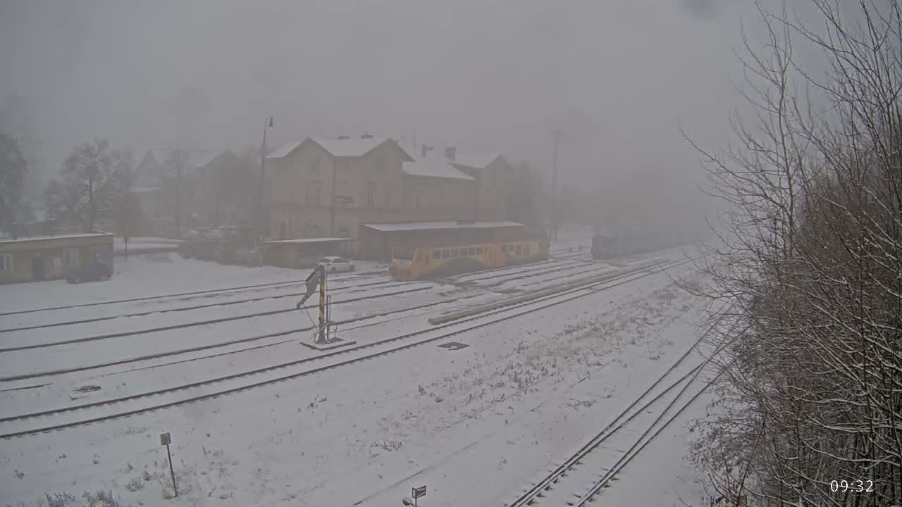 A snow-covered train station with multiple tracks, a parked yellow and green train, and bare trees is shrouded in heavy fog and falling snow.