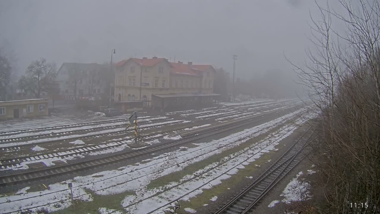 Multiple snow-dusted railway tracks lead towards a prominent station building and other structures in the distance, all heavily obscured by dense fog on a cold, overcast day.