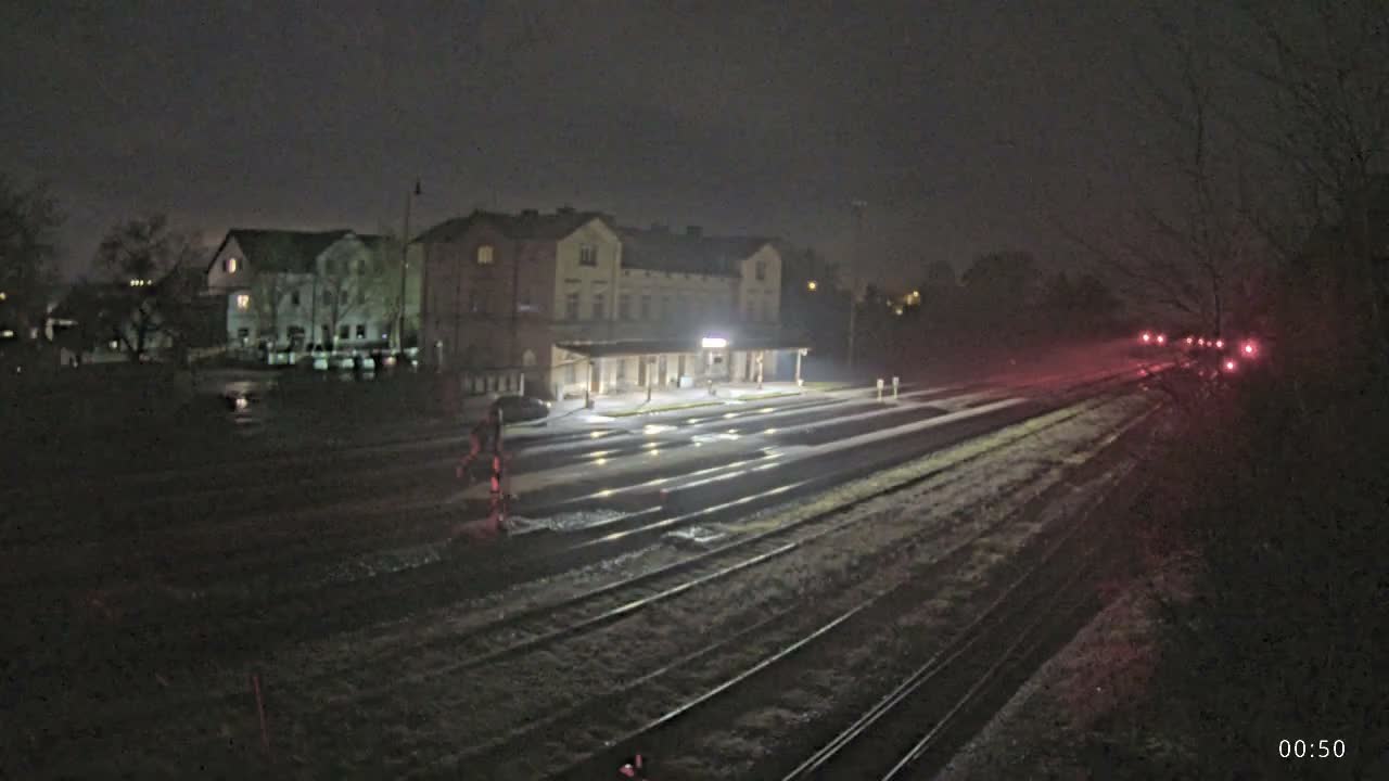 A nighttime view reveals a train station with multiple wet tracks reflecting artificial lights, an illuminated main building with a covered platform, and distant red signals, all under dark and damp conditions.
