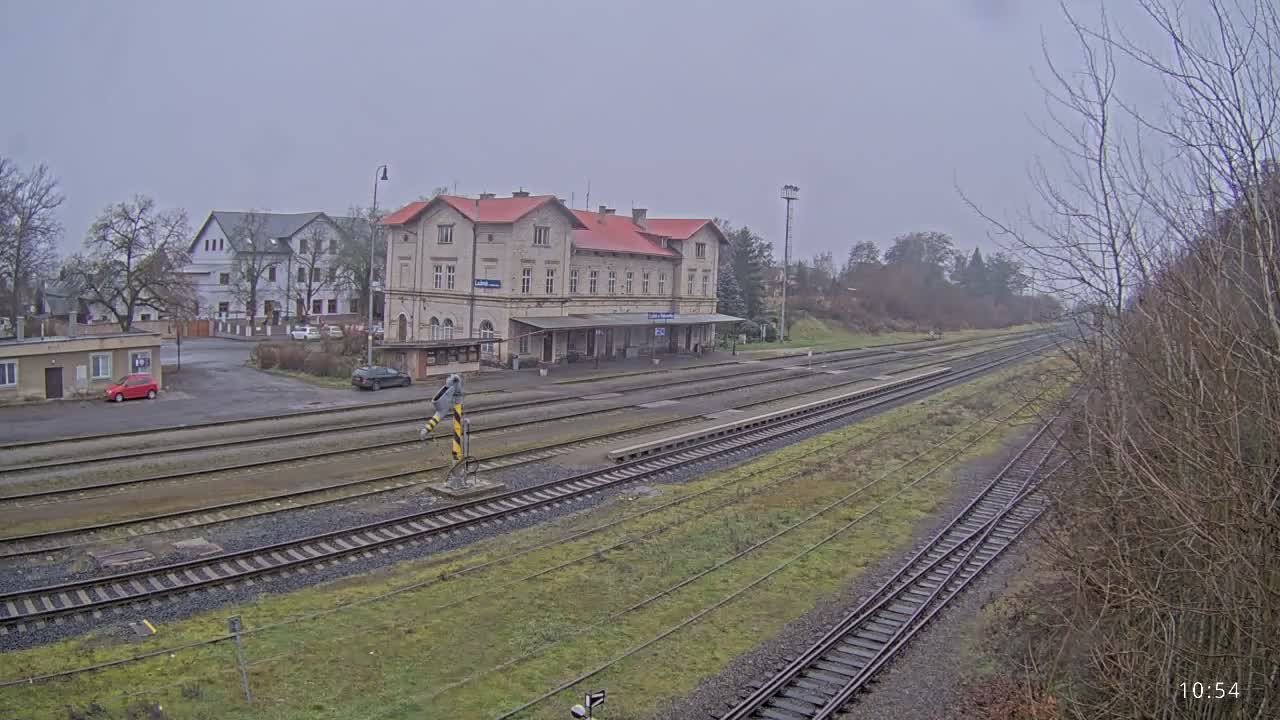 A nighttime view reveals a train station with multiple wet tracks reflecting artificial lights, an illuminated main building with a covered platform, and distant red signals, all under dark and damp conditions.