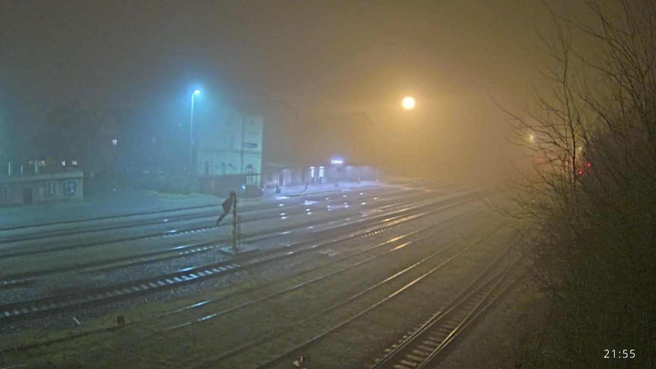 A nighttime view reveals a train station with multiple wet tracks reflecting artificial lights, an illuminated main building with a covered platform, and distant red signals, all under dark and damp conditions.