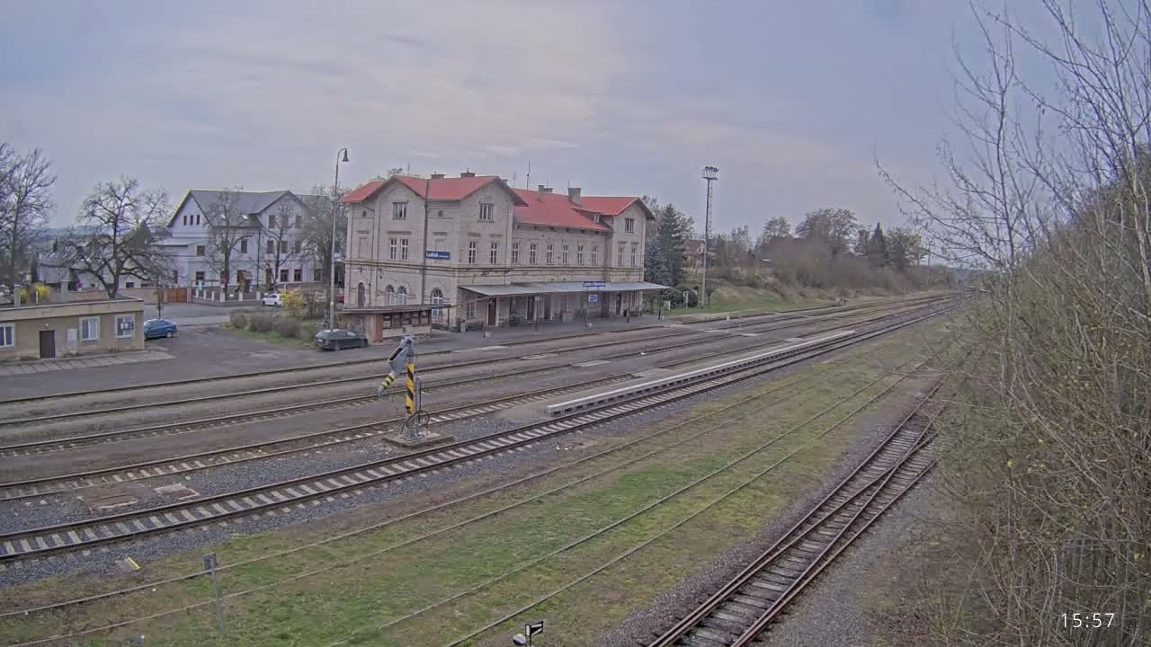 A nighttime view reveals a train station with multiple wet tracks reflecting artificial lights, an illuminated main building with a covered platform, and distant red signals, all under dark and damp conditions.