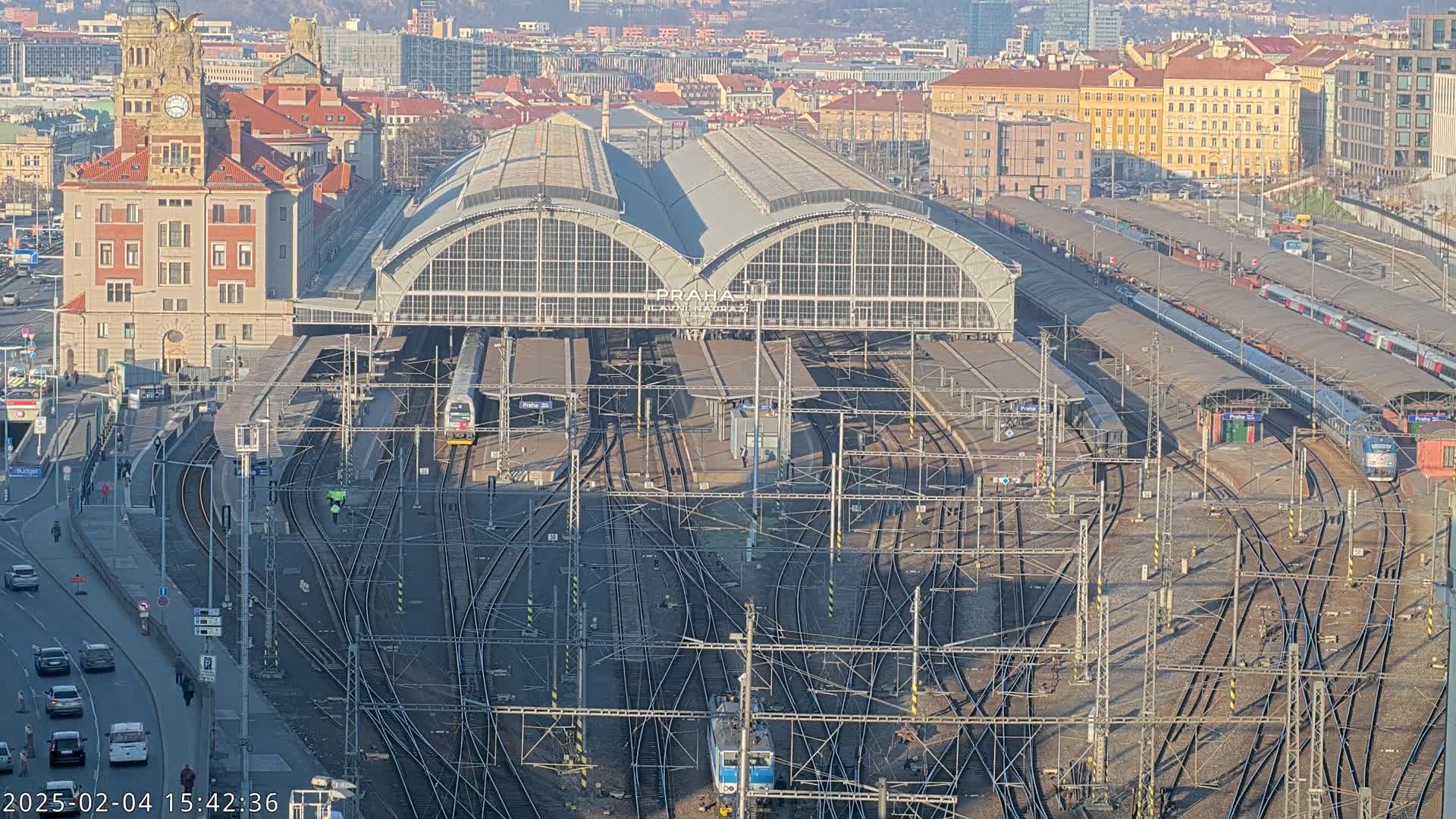 A large train station with numerous tracks and trains, surrounded by city buildings under a bright, sunny sky.