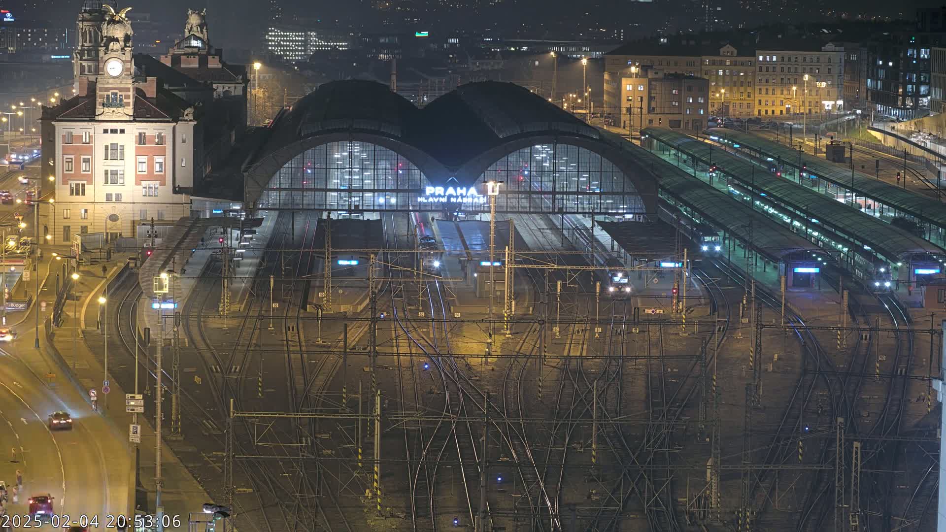 A nighttime aerial view of a large train station with numerous tracks and platforms, surrounded by city buildings and illuminated roads.