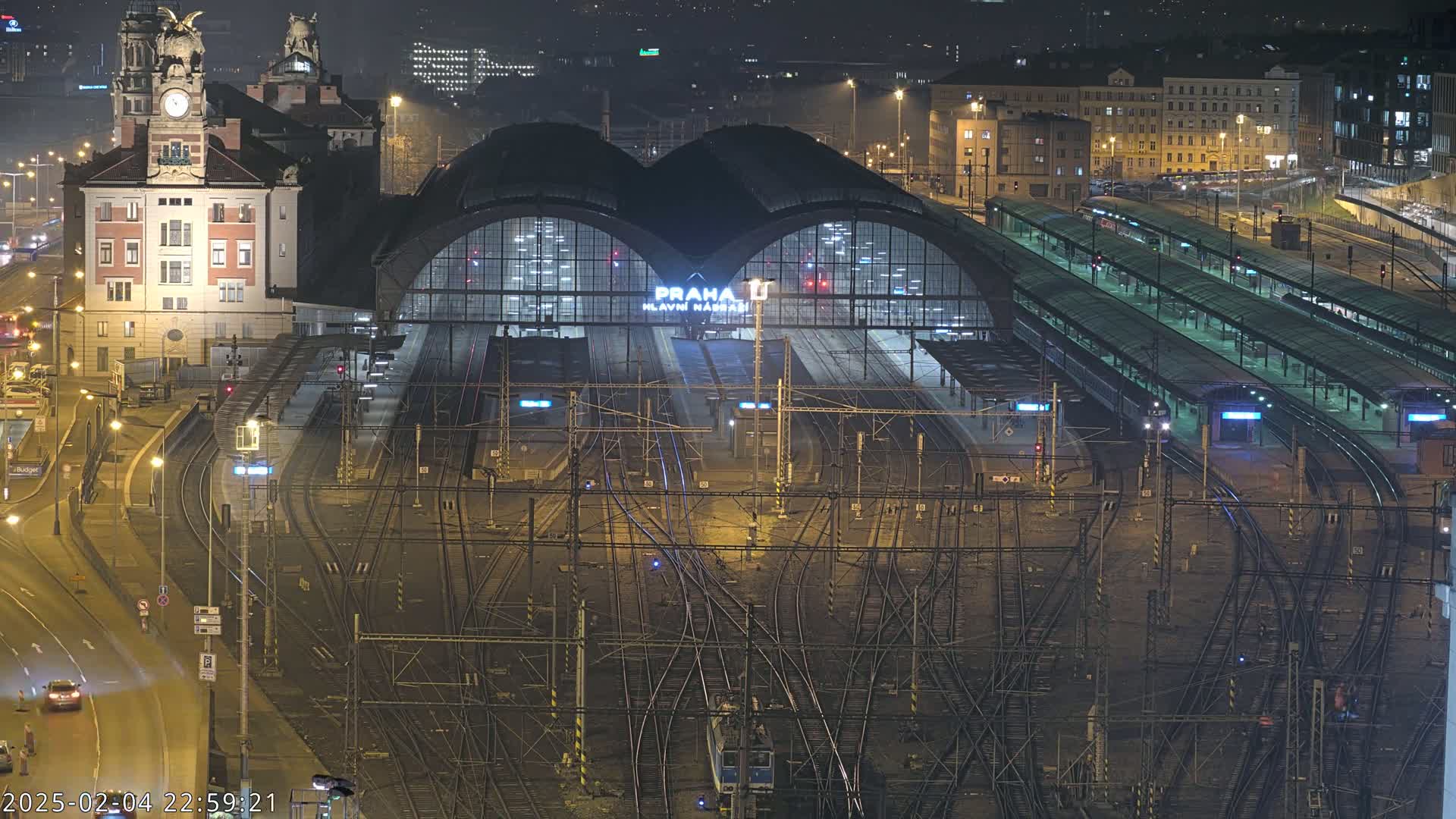 A large, illuminated train station at night, with numerous train tracks and platforms extending from a central building, under a dark sky.