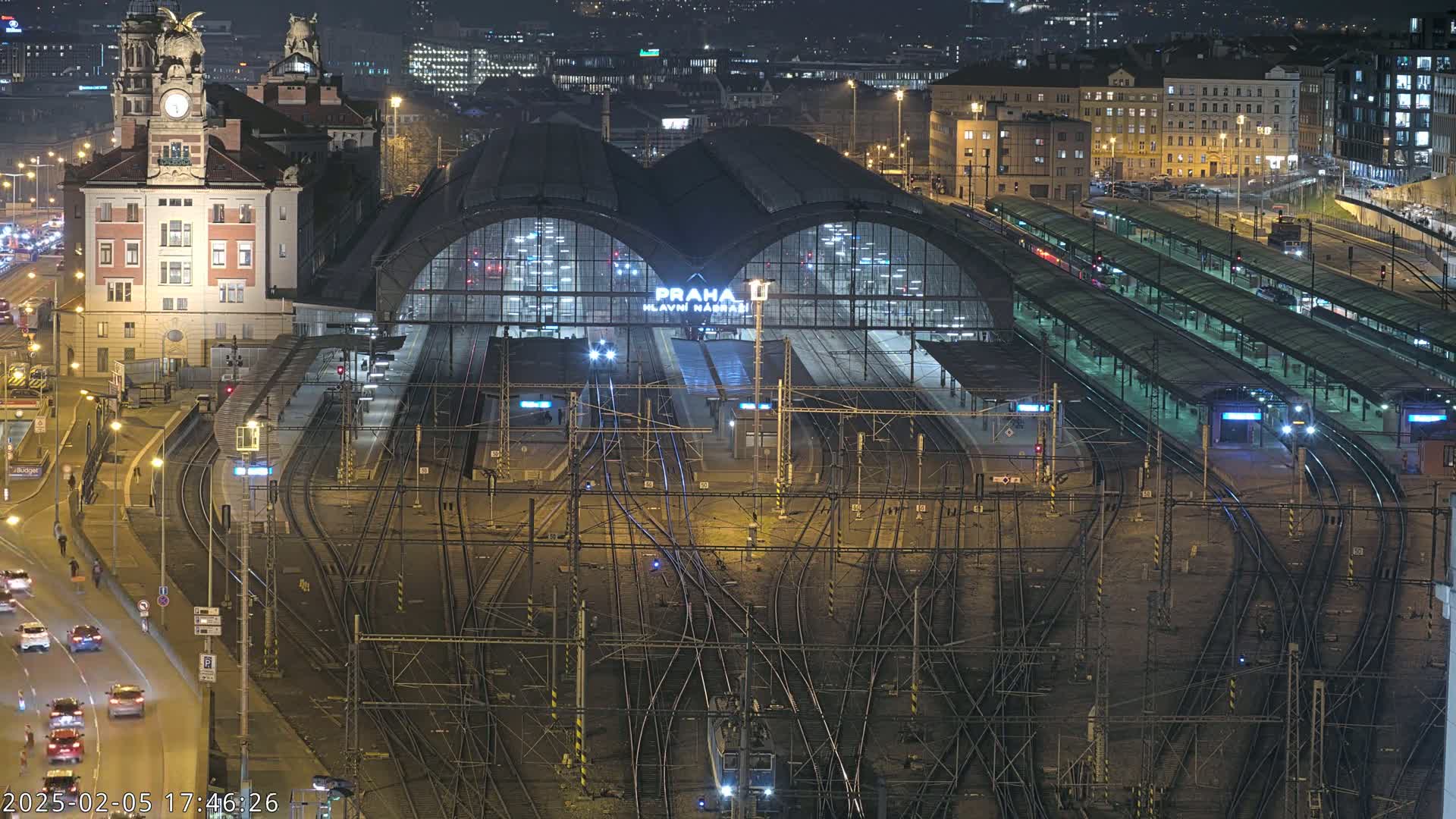 A brightly lit, large train station at night with many tracks and trains, surrounded by city buildings under a dark sky.