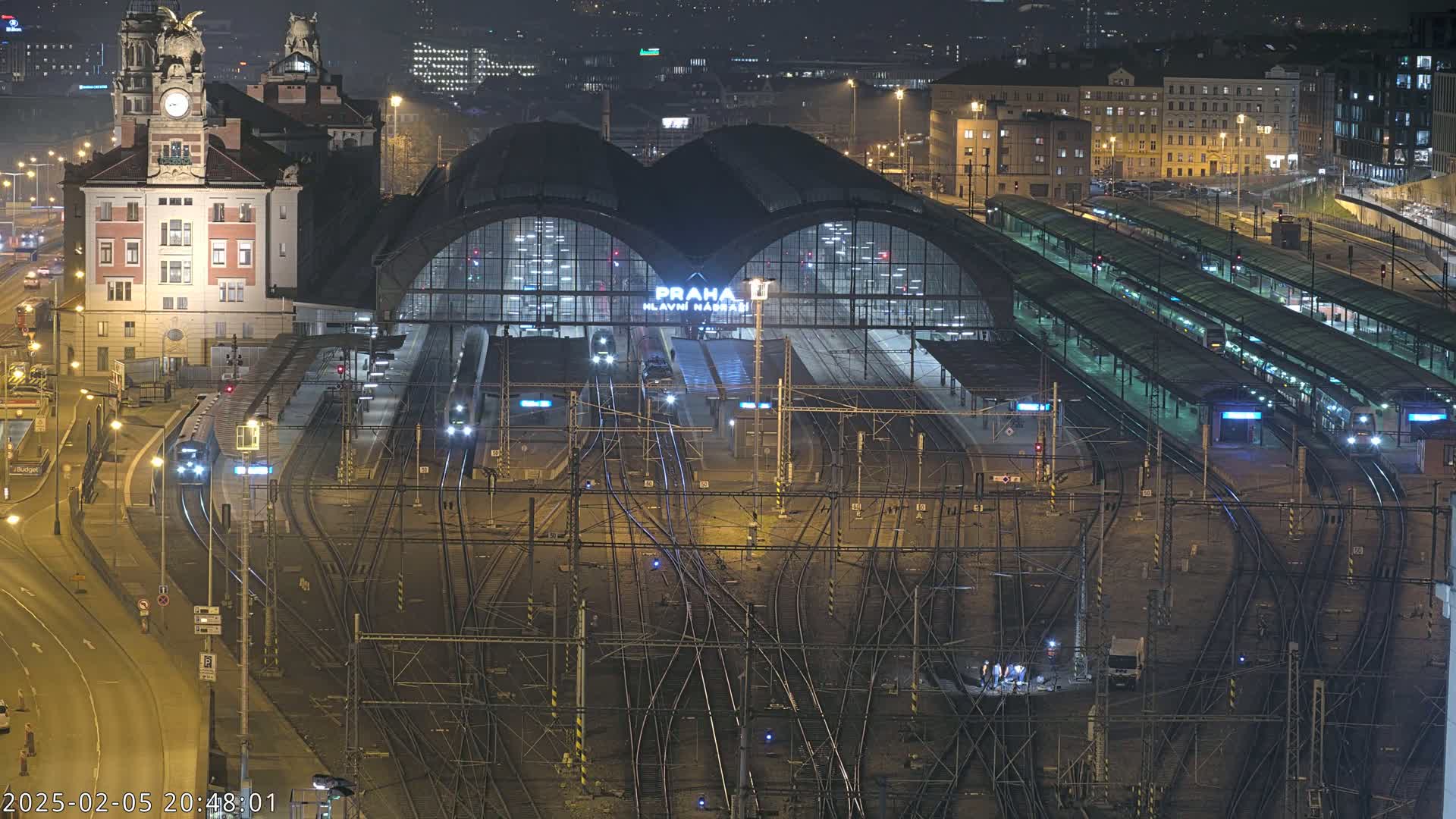 A nighttime, high-angle view of a busy train station with numerous tracks, platforms, and trains, surrounded by city buildings.