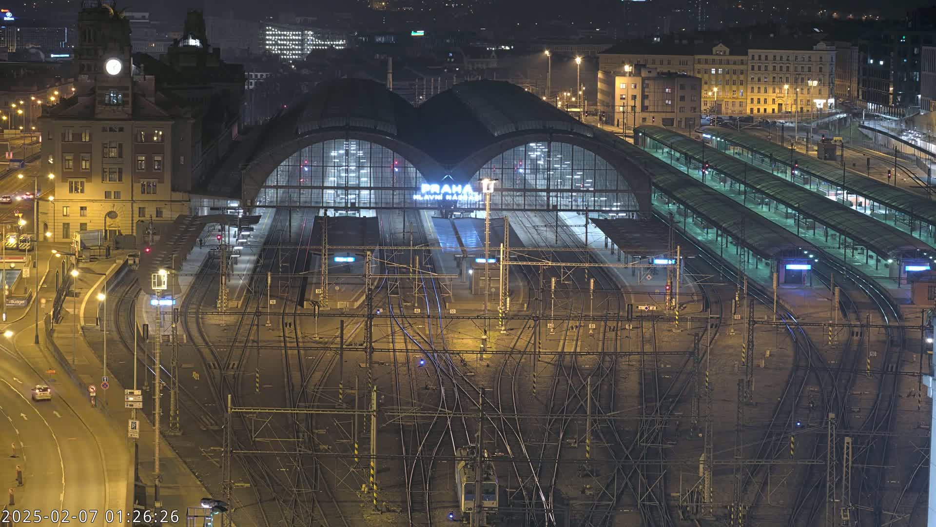A large, illuminated train station at night, surrounded by city buildings and a complex network of train tracks.