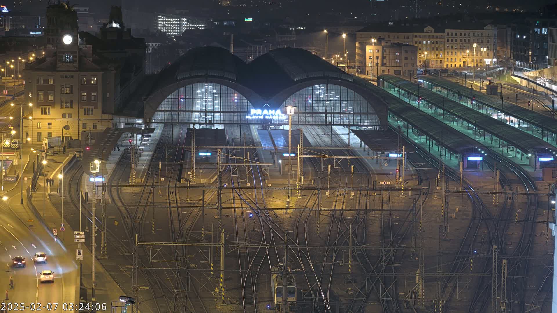 A nighttime, aerial view of a large, illuminated train station with numerous tracks and platforms, surrounded by city buildings.