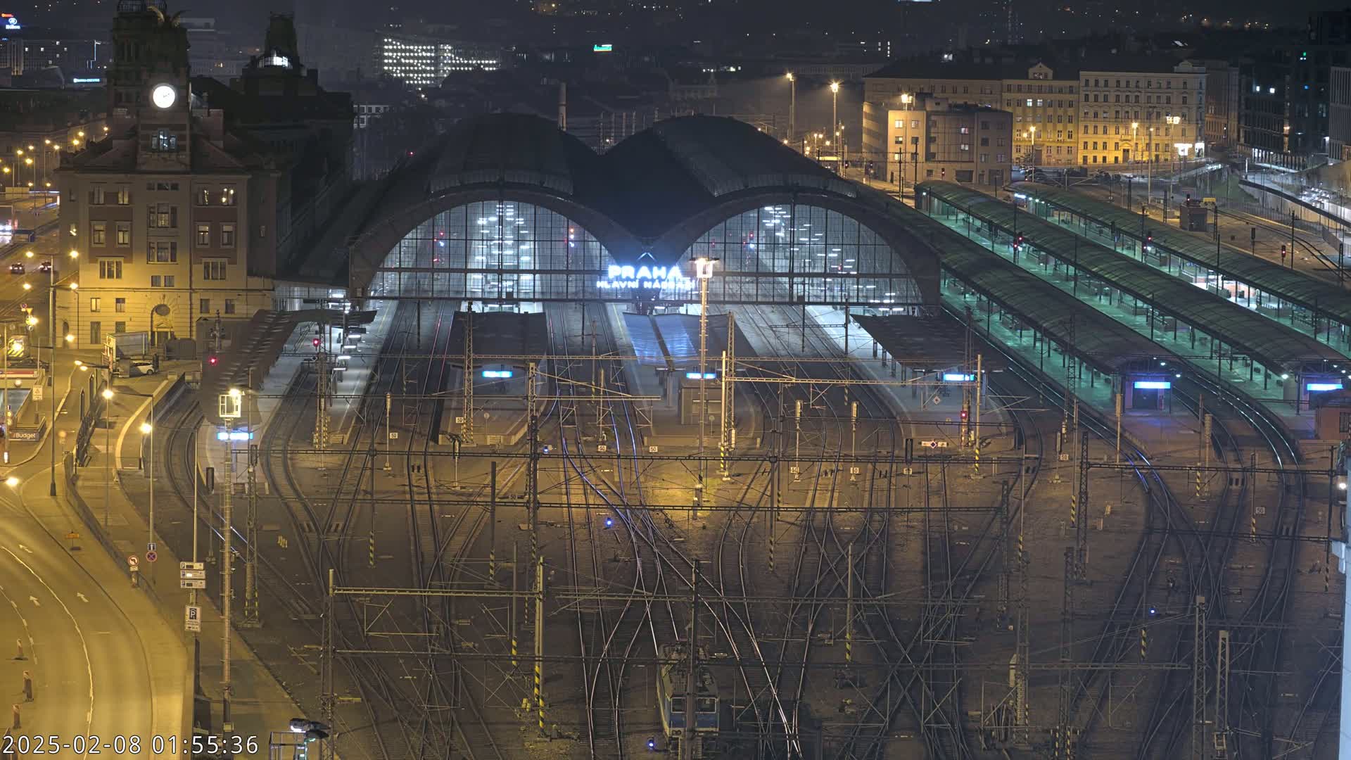 A large, illuminated train station at night, with many tracks and platforms extending from it, surrounded by city buildings.