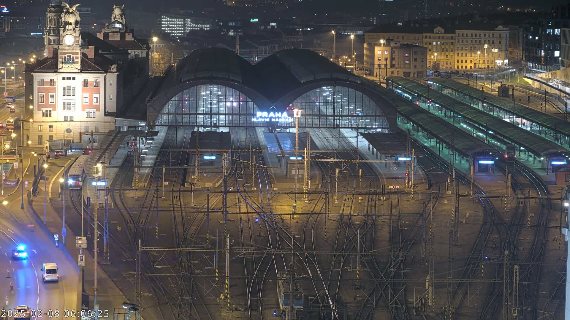 A nighttime aerial view of a large, illuminated train station with numerous tracks and platforms, surrounded by city buildings.