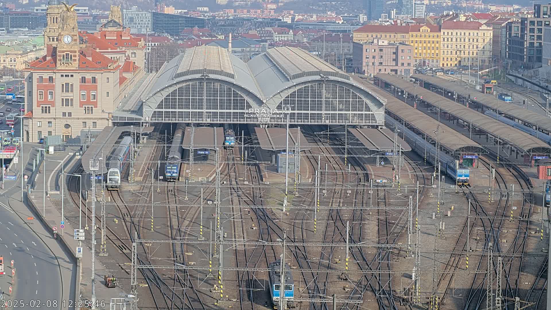 A large train station with numerous tracks and trains, under a sunny sky, is situated amidst a city.