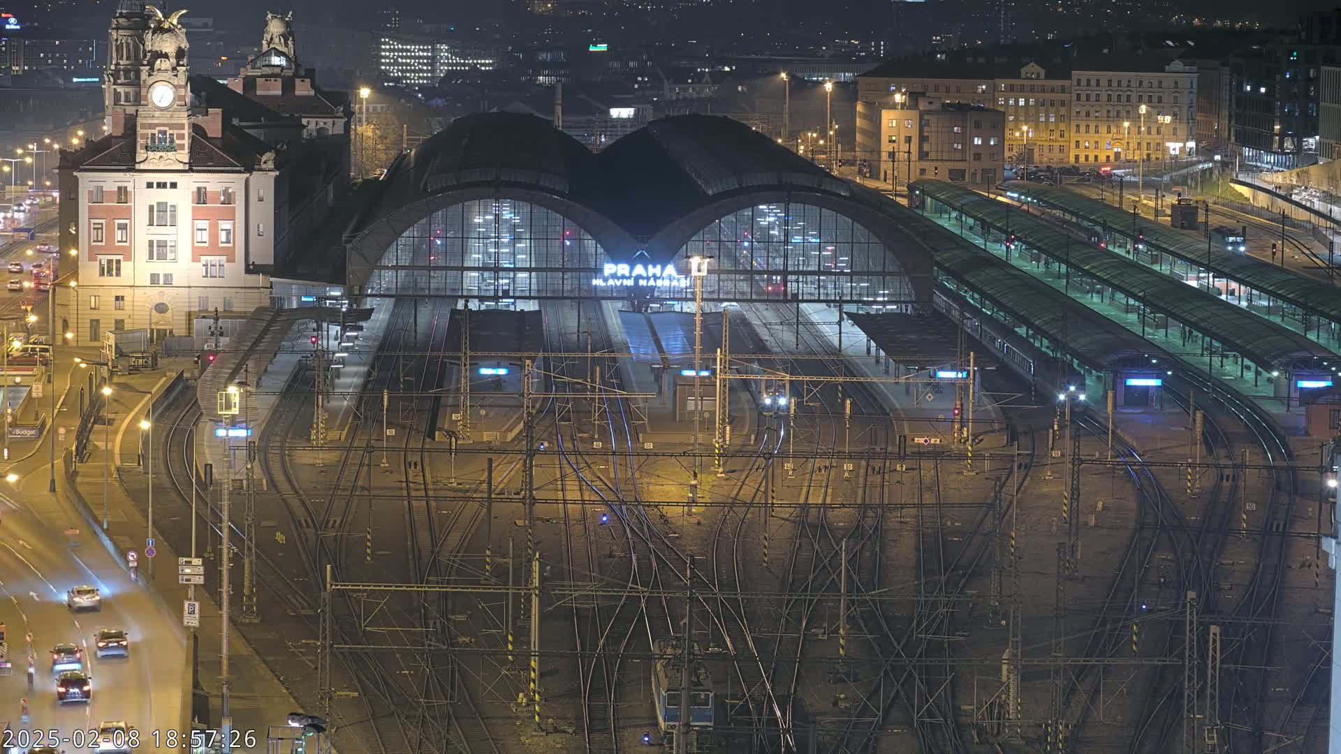 A large, illuminated train station at night, with numerous tracks and platforms extending from it, surrounded by city buildings and roads with cars.