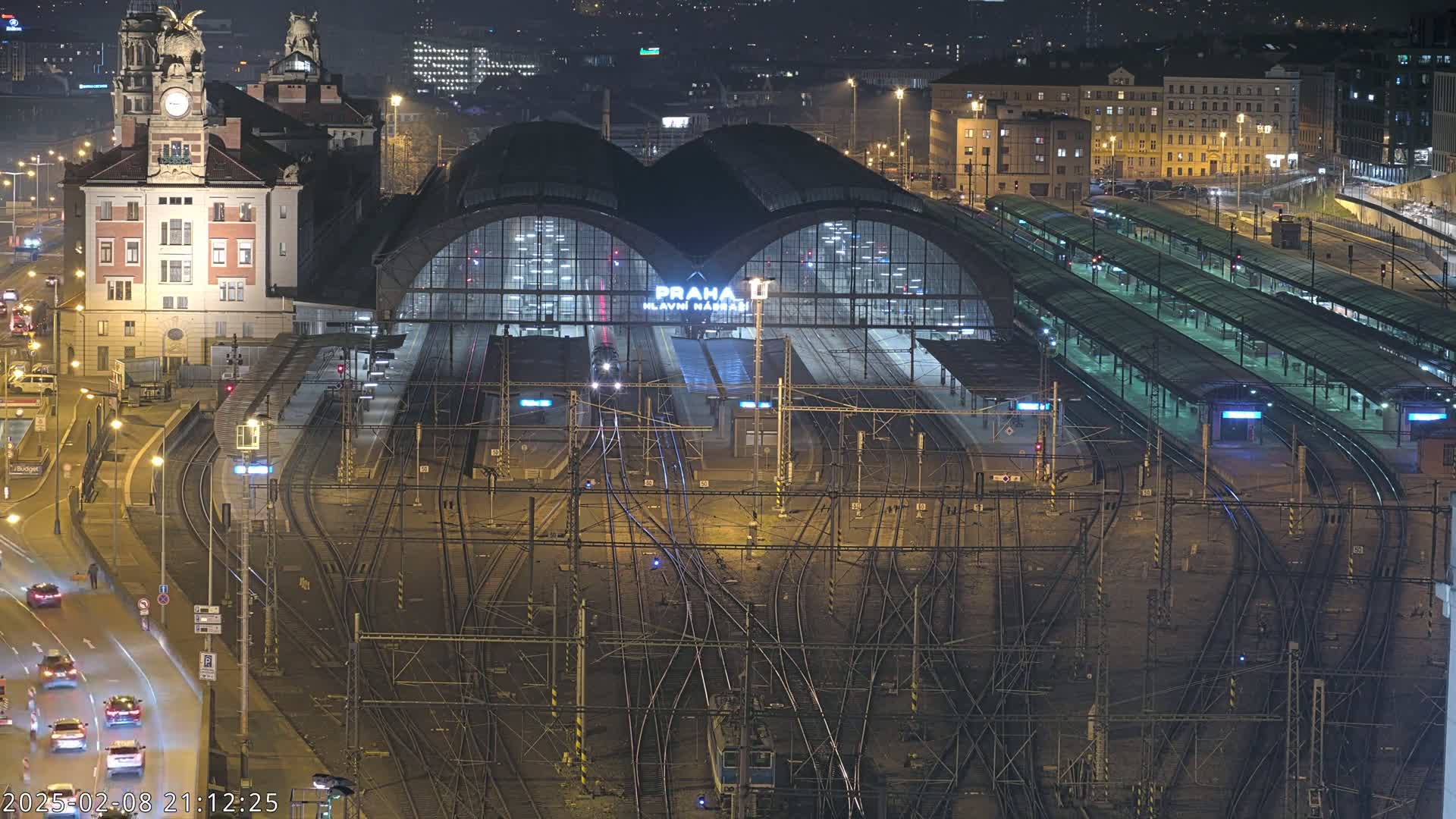 A nighttime aerial view of a large, illuminated train station with numerous tracks and platforms, surrounded by city buildings.