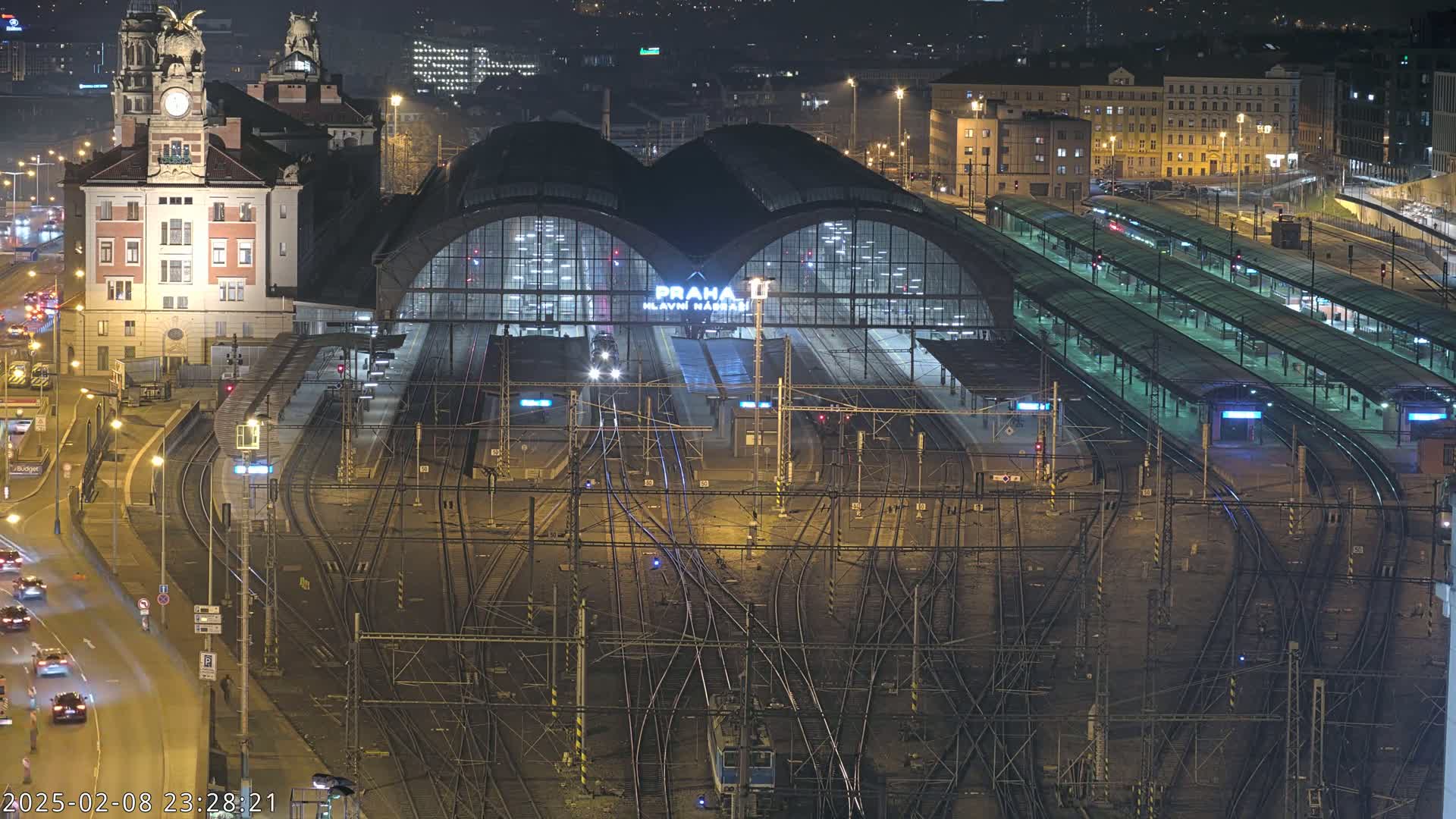 A nighttime, aerial view of a large, illuminated train station with numerous tracks and platforms, surrounded by city buildings.