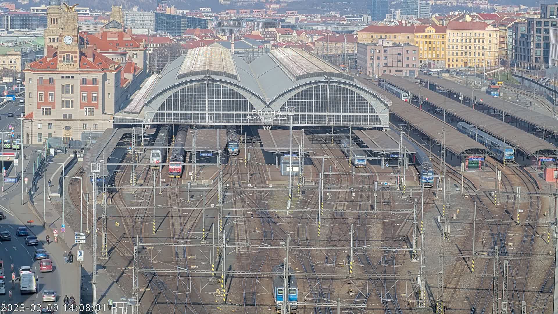 A large train station with many tracks and trains, under a sunny sky, is surrounded by city buildings.