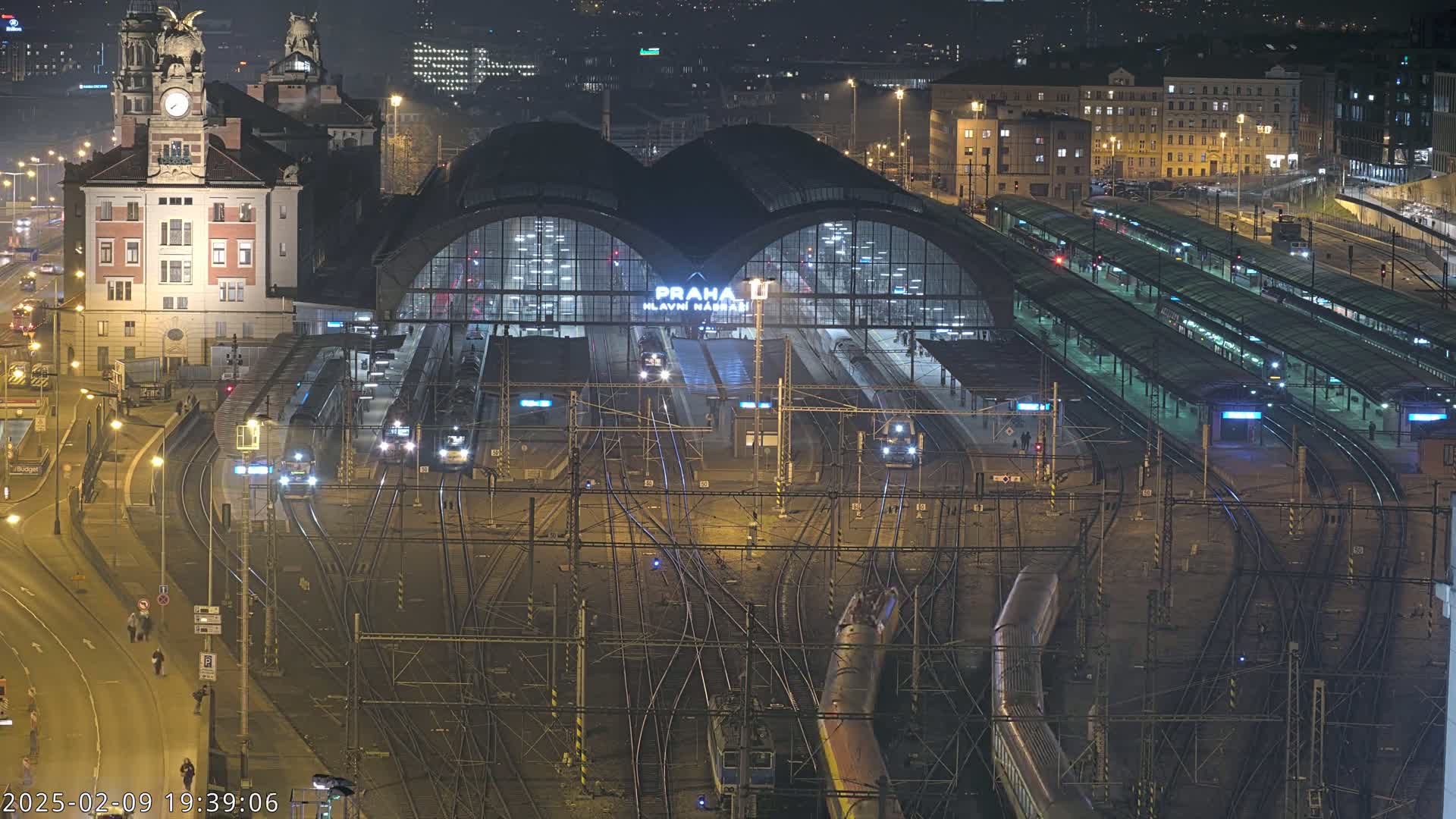 A nighttime, high-angle view of a large, busy train station with numerous tracks and trains, surrounded by city buildings.