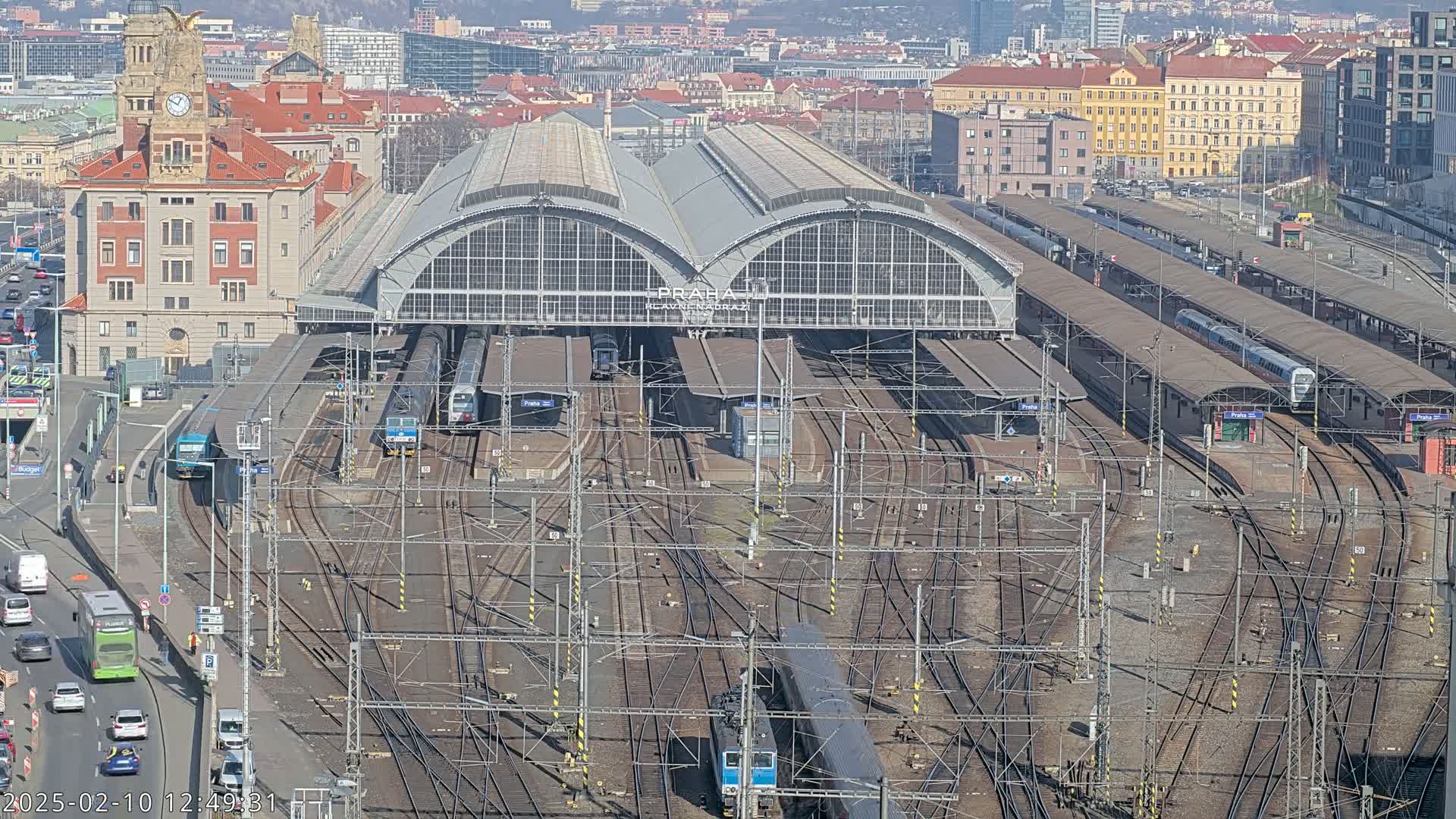 A large train station with numerous tracks and trains, surrounded by city buildings under a sunny sky.
