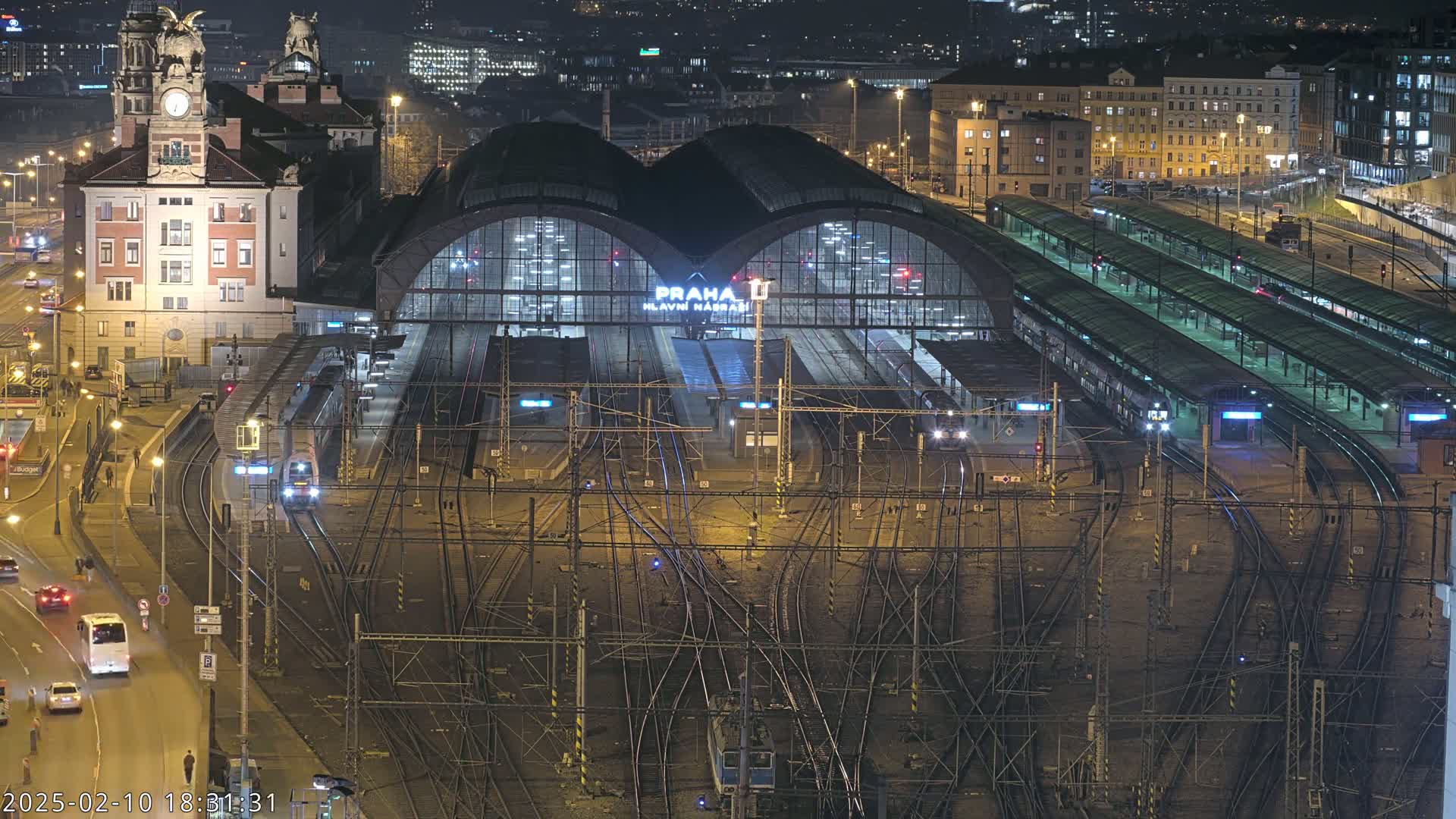 A brightly lit train station at night, with many trains and tracks visible, surrounded by city buildings and roads.
