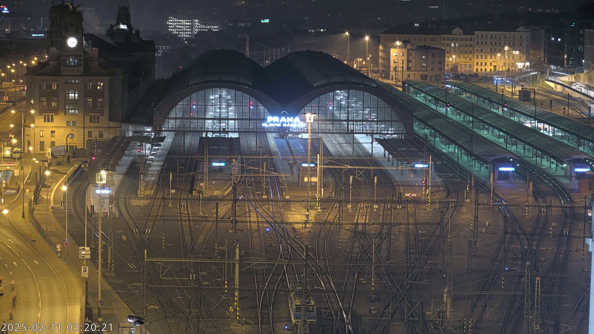A nighttime aerial view of a large, illuminated train station with numerous tracks and platforms, surrounded by city buildings.