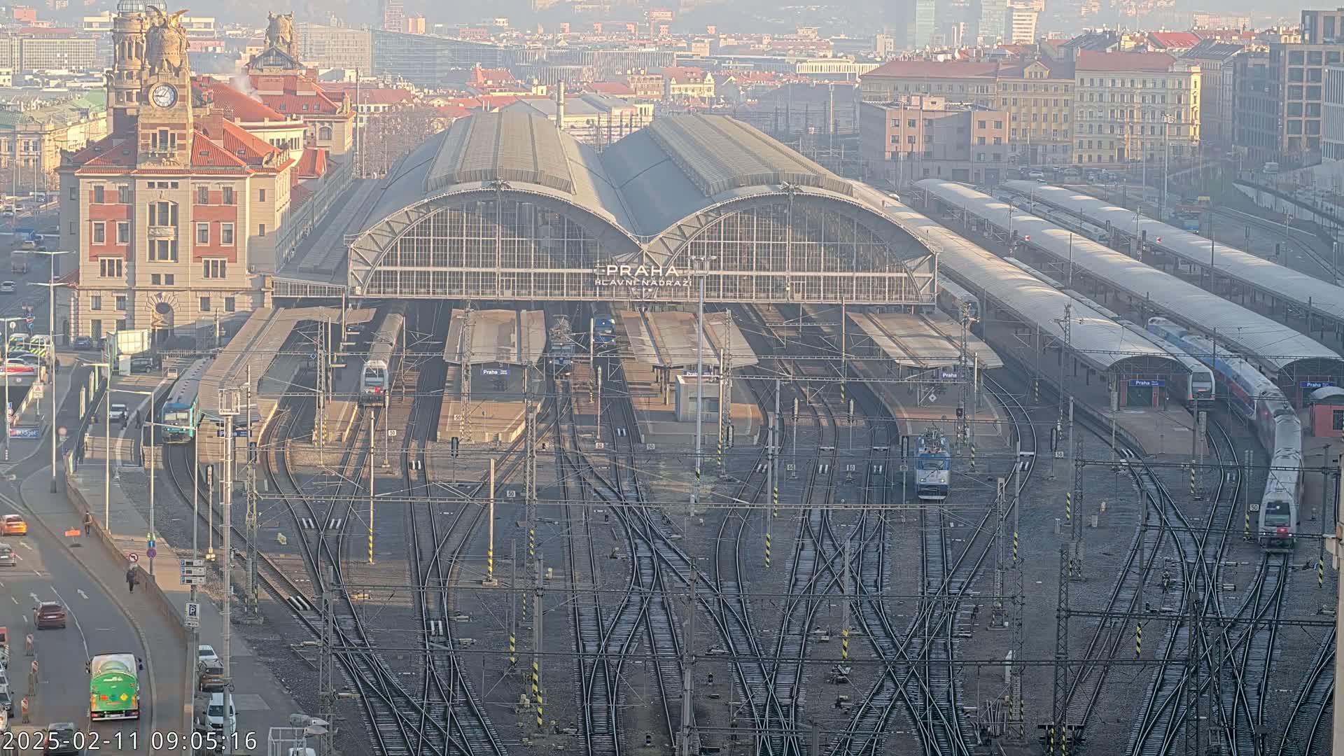 A large train station with numerous tracks and trains, surrounded by city buildings under a hazy, sunny sky.