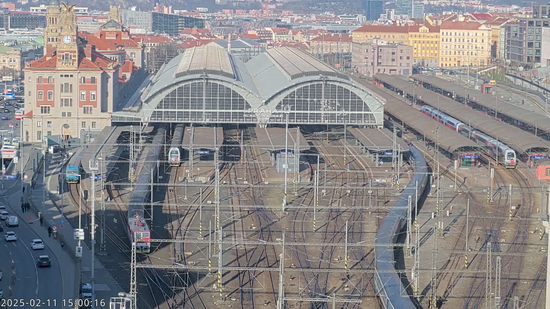 A large train station with numerous trains at platforms under a sunny sky, surrounded by city buildings.