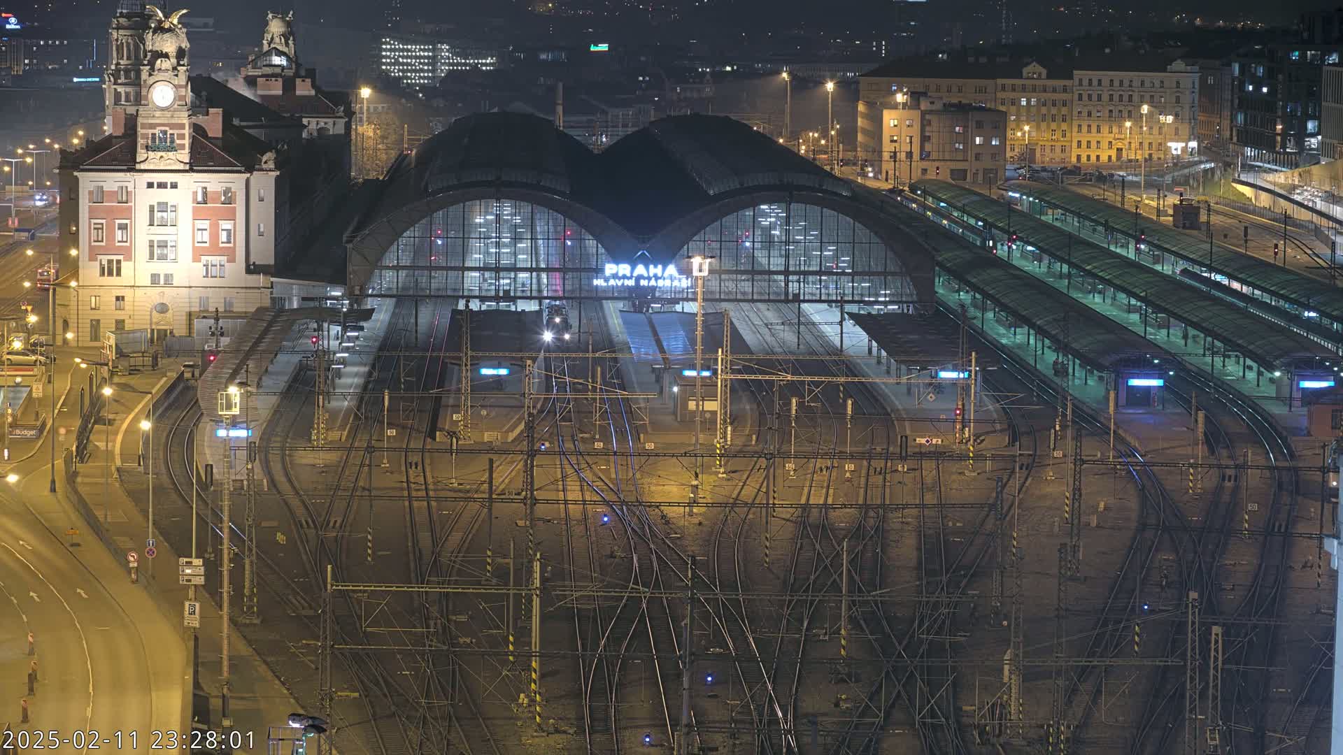 A large train station at night, illuminated, with many tracks and platforms, and a tall building adjacent to it.