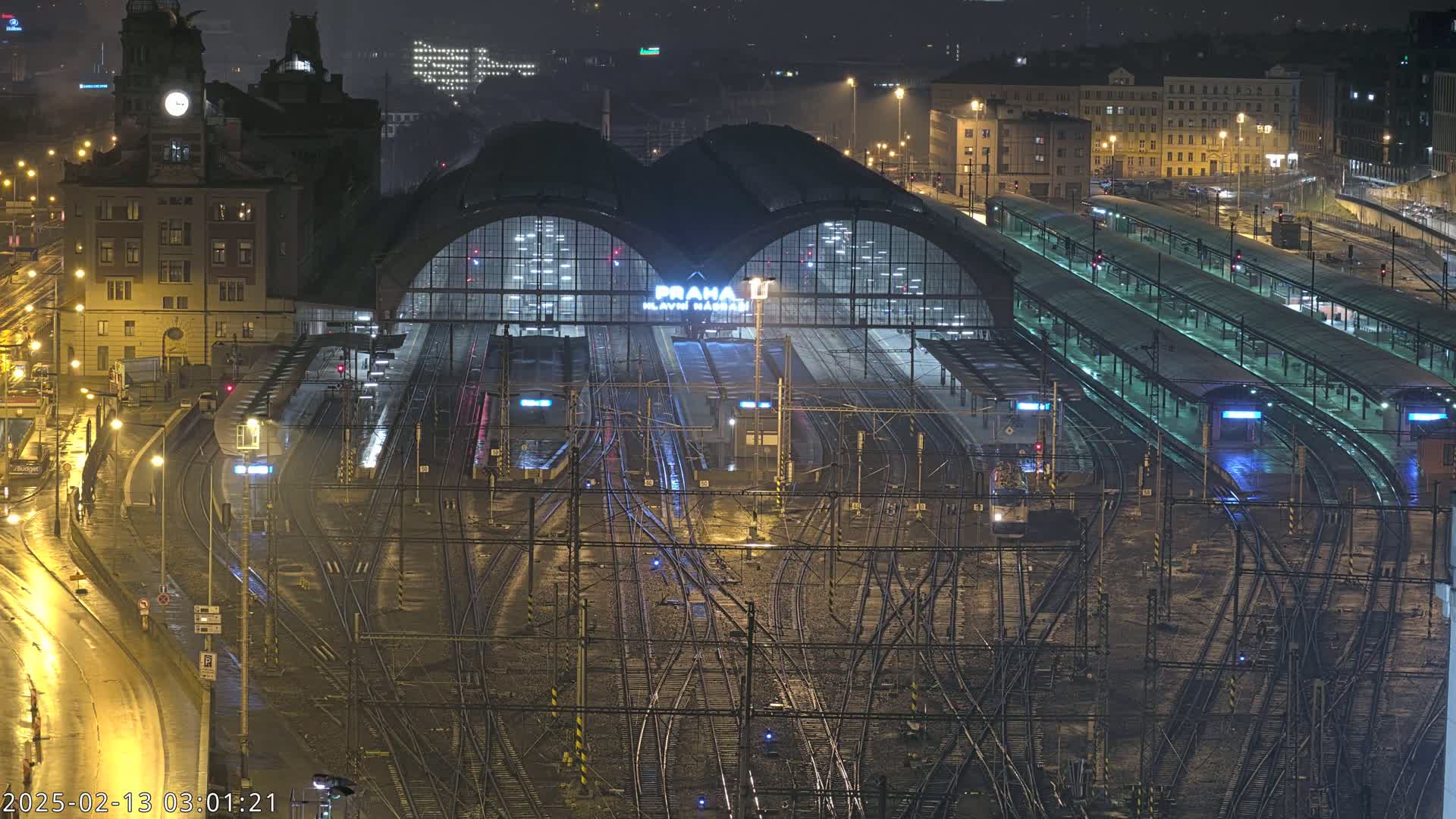 A nighttime, wet view of a large train station with many tracks and trains, surrounded by city buildings.