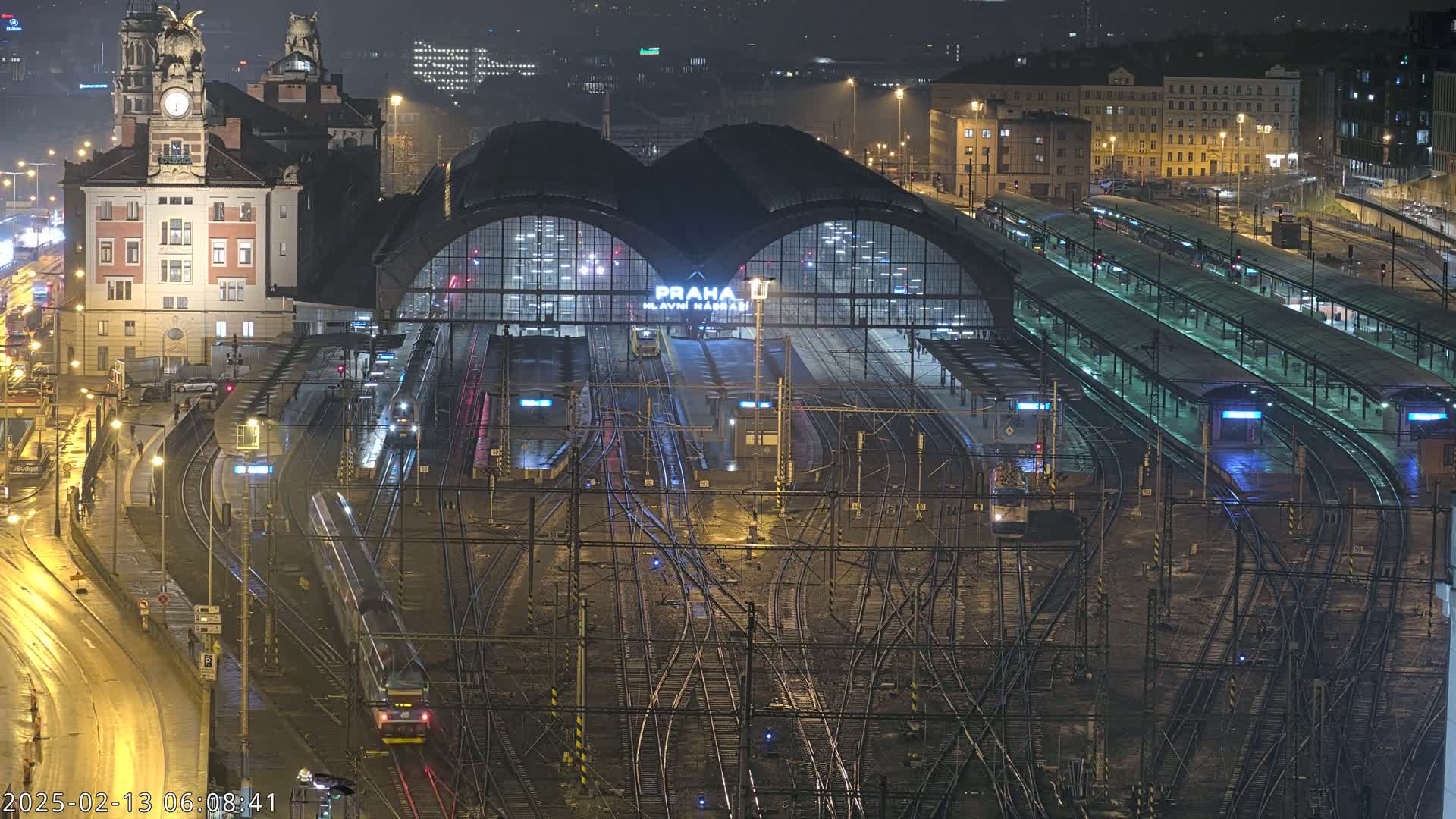 A nighttime view of a large, illuminated train station with numerous tracks and trains, wet roads, and surrounding city buildings under a dark sky.