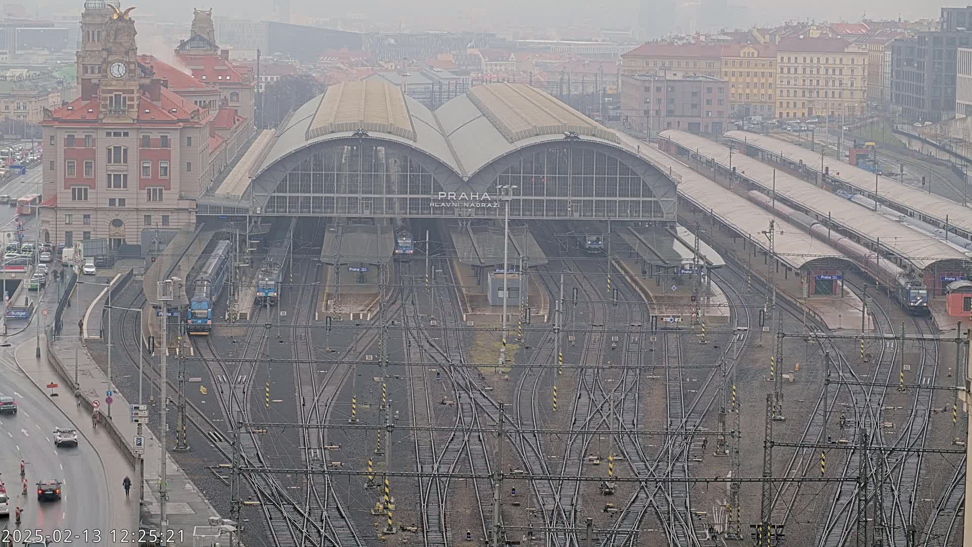 A large train station with numerous tracks and trains, under an overcast sky with light rain.