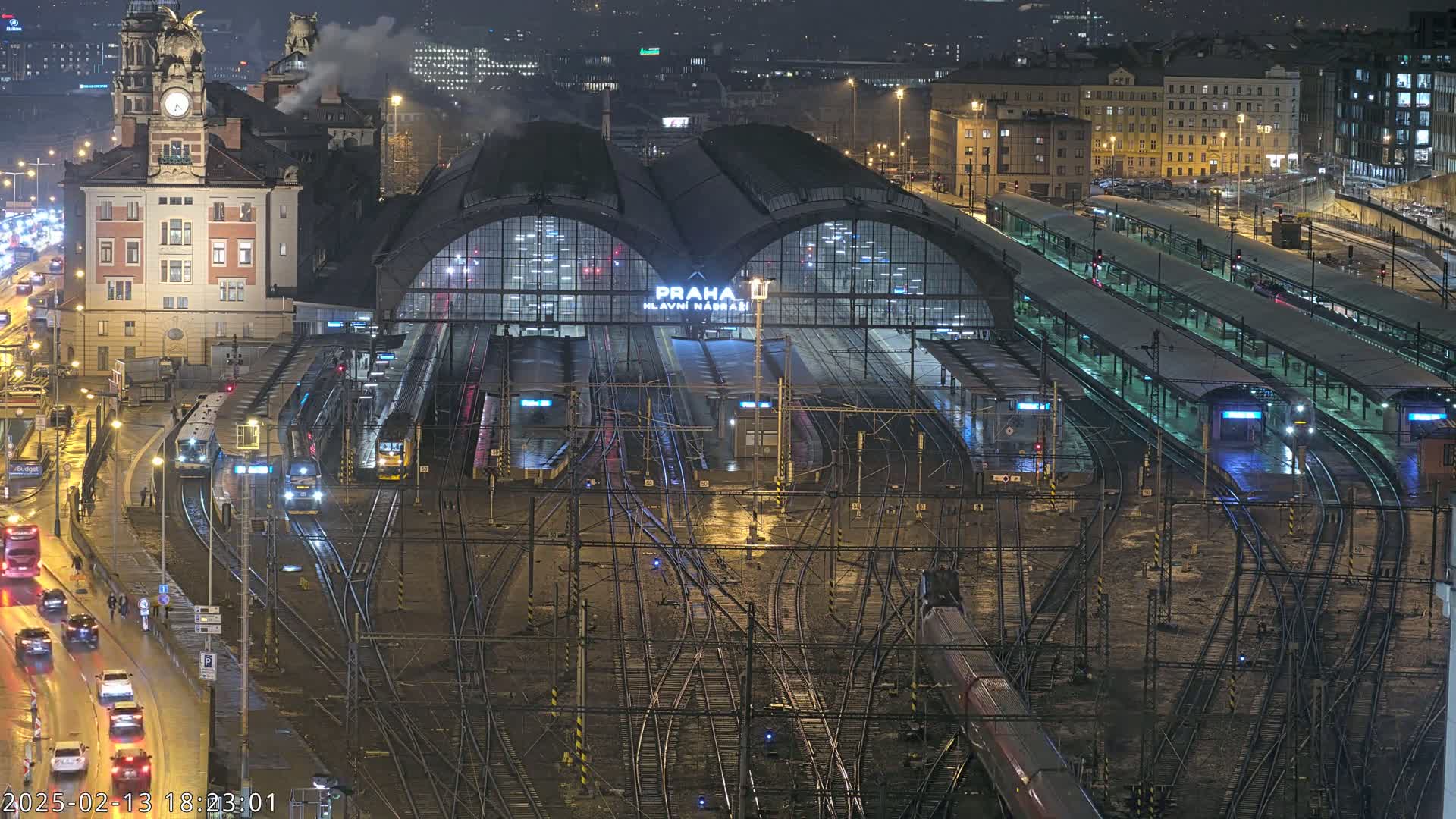 A nighttime view of a large, busy train station with numerous tracks and trains, surrounded by city buildings under wet conditions.