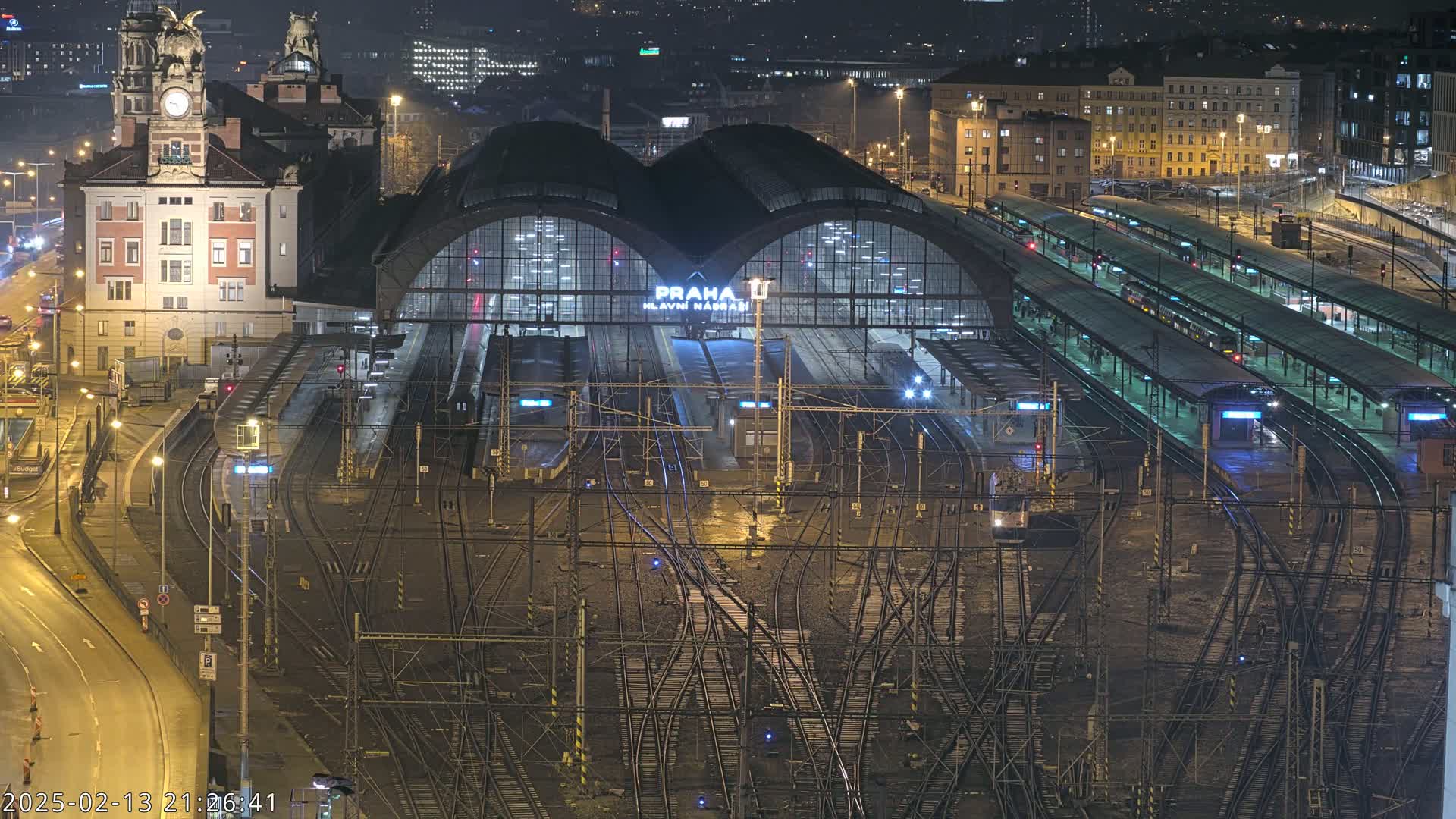 A nighttime aerial view of a large, illuminated train station with numerous tracks and trains, surrounded by city buildings.