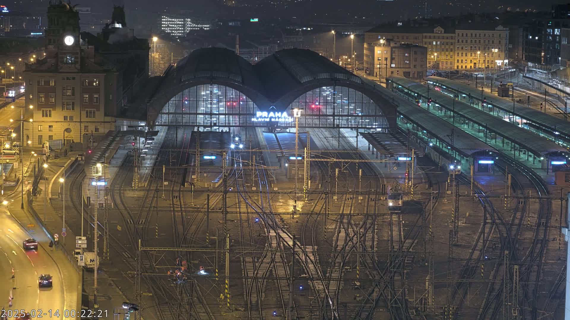 A nighttime aerial view of a large, illuminated train station with many tracks and platforms, surrounded by city buildings.