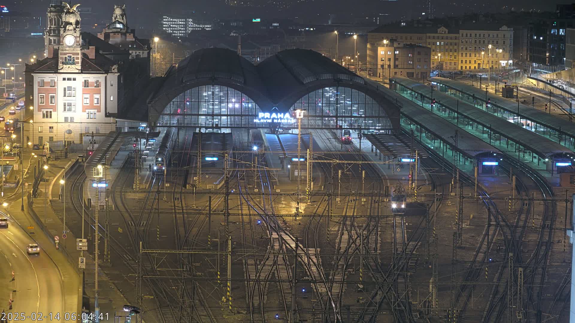 A nighttime, aerial view of a large train station with numerous tracks and platforms, surrounded by city buildings.