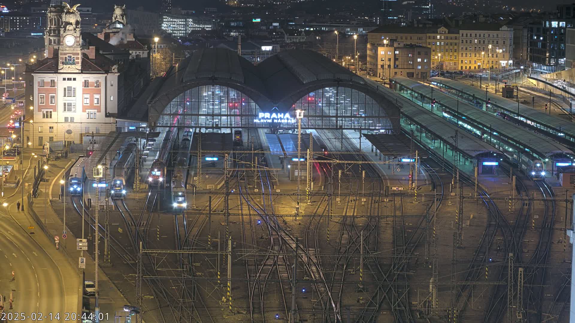 A nighttime aerial view of a large train station with numerous trains and tracks, surrounded by city buildings.