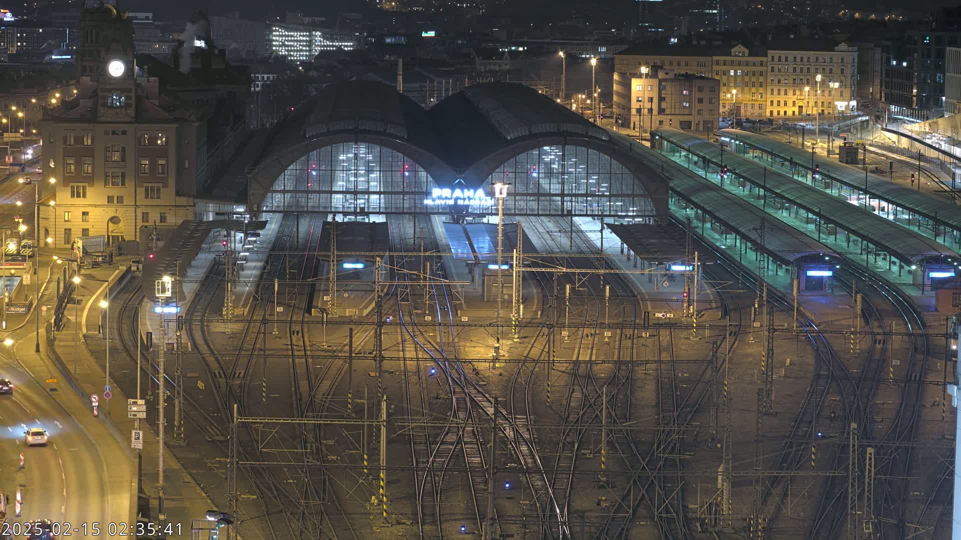 A large, illuminated train station at night, with numerous train tracks and platforms extending from it, surrounded by city buildings under a dark sky.